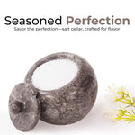 A white marble salt cellar with lid containing pink salt, placed on a white surface with some plant parts in the background.