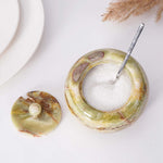 A white marble salt cellar with lid containing pink salt, placed on a white surface with some plant parts in the background.