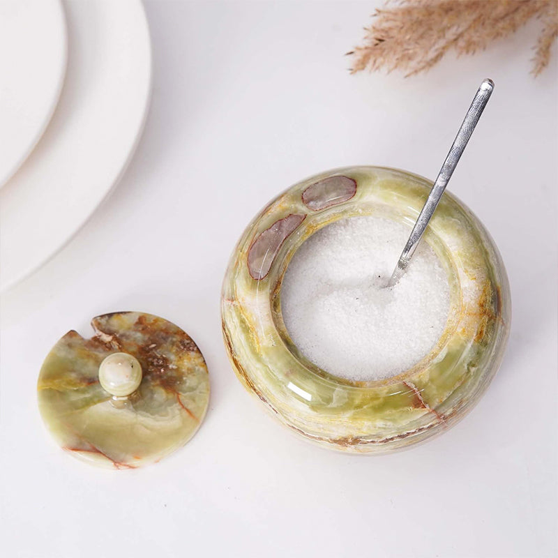 A white marble salt cellar with lid containing pink salt, placed on a white surface with some plant parts in the background.