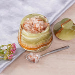 Decorative container with pink Himalayan salt, spoon, and flowers on a light surface