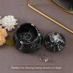 Black marble jewelry box with a ring on a gray surface with flowers and a mirror.