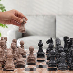 A person's hand lifting a white marble chess piece off the board, with other pieces arranged for play. The board is on a marble-like surface with a patterned background.
