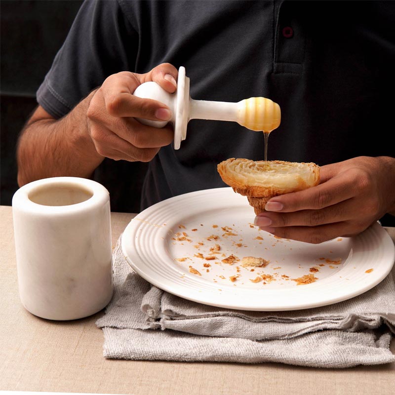 A person is pouring honey from a white marble honey jar into a small white bowl, which is placed on a white plate with a cup and some bread.