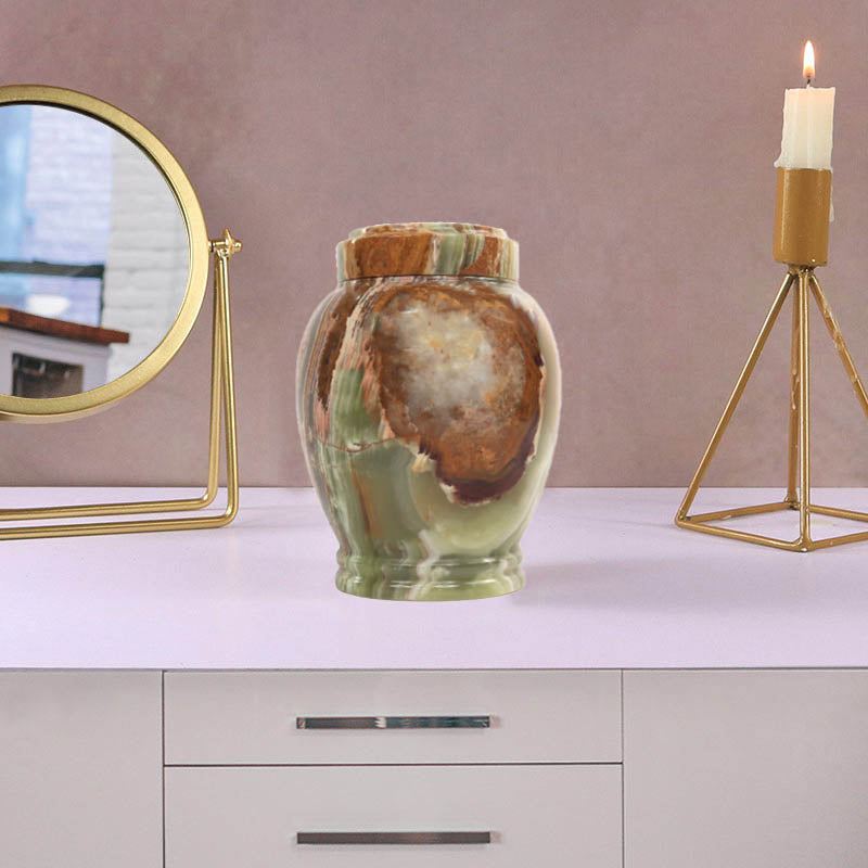 A marble classic urn with a green and brown pattern, placed on a white surface, accompanied by a golden framed mirror and a candle holder in the background.