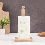 A white square towel holder made of onyx marble is displayed with a roll of paper towels partially unwound. It is placed on a table with a blue vase and decorative flowers in the background, alongside a pair of sunglasses.