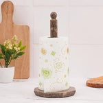 A white paper towel roll is placed on a onyx marble towel holder, which is itself supported by a circular base. This setup is positioned on a table with a vase of flowers and a cup in the background.