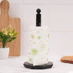 A white paper towel roll is placed on a onyx marble towel holder, which is itself supported by a circular base. This setup is positioned on a table with a vase of flowers and a cup in the background.