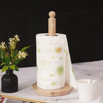A white paper towel roll is placed on a onyx marble towel holder, which is itself supported by a circular base. This setup is positioned on a table with a vase of flowers and a cup in the background.