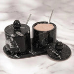 A black marble salt cellar with an oval tray and two stainless steel spoons, placed on a marble countertop.