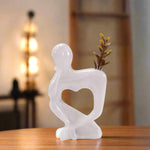 A white marble vase in the shape of a lady's torso with a heart-shaped cutout for flowers, placed on a wooden surface in a home environment.