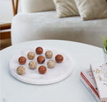 A game of Tic Tac Toe with marble balls on a white board, placed on a white table with a couch and a book in the background.