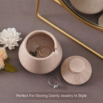 Beige jewelry dish with earrings and ring on a gray surface with flowers and mirror.