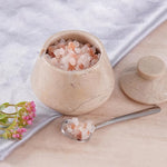 Marble container with pink Himalayan salt, spoon, and flowers on a light surface.