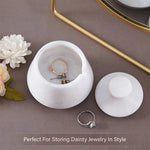 White marble jewelry box with lid open, displaying gold and silver rings on a brown surface with flowers and a mirror.