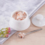 White ceramic bowl filled with pink Himalayan salt on a light wooden surface with a spoon and small plant.