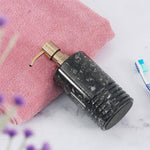 A black marble soap dispenser with a gold pump, placed on a white surface next to a pink towel and a toothbrush.