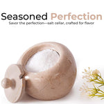 A white marble salt cellar with lid containing pink salt, placed on a white surface with some plant parts in the background.