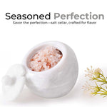 A white marble salt cellar with lid containing pink salt, placed on a white surface with some plant parts in the background.