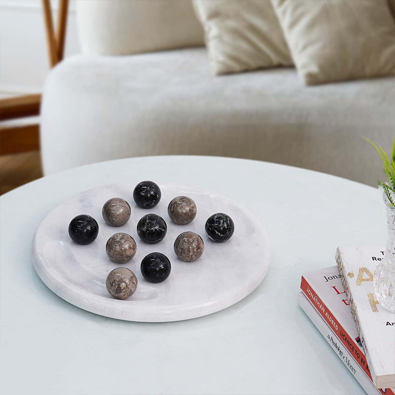 A Tic Tac Toe board with black and white marble balls on a white surface, with a couch and a plant in the background.